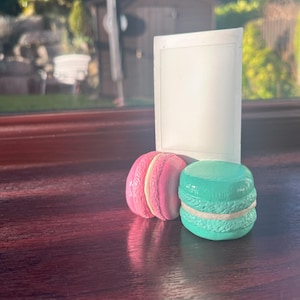 May include: Two macarons, one pink and one teal, sit in front of a blank white sign. The macarons have a white filling.