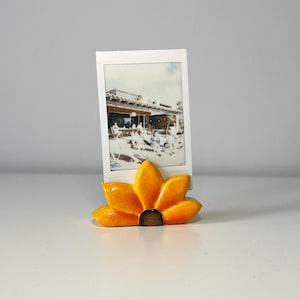 May include: A ceramic photo holder in the shape of a sunflower. The holder is bright yellow with a brown center and holds a white framed photo. The photo shows a group of people outside a building.