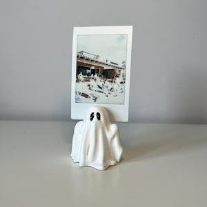 Op de afbeelding: Een witte keramische fotohouder in de vorm van een geest met een glanzende afwerking en twee zwart geschilderde ogen. Het geestfiguur heeft een gleuf aan de bovenkant die een rechthoekige instantfoto van een buitenterras stevig vasthoudt.