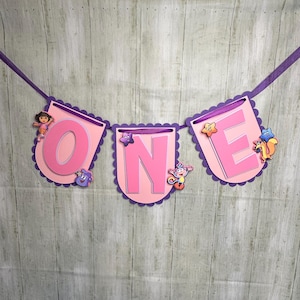 May include: A decorative party banner featuring three scalloped-edge panels in light pink and purple, spelling out the word ONE in large pink letters. The banner is strung on a purple ribbon and decorated with cartoon character cutouts.