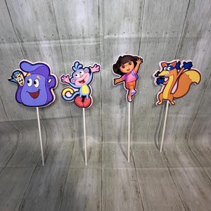 May include: Four colorful character cutouts on white sticks. The characters include a purple backpack with a smiling face, a blue monkey, a girl in a pink shirt and orange pants, and an orange fox with a blue mask. These are likely cake toppers or party decorations.