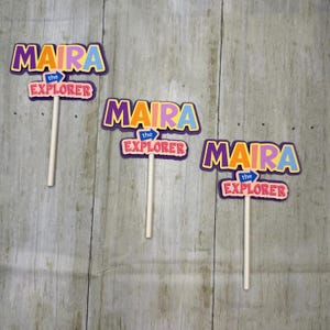 Puede incluir: Tres adornos decorativos para cupcakes con el texto MAIRA THE EXPLORER en letras de bloque coloridas. Cada adorno tiene un palito blanco en la parte inferior. La palabra MAIRA es amarilla, azul y morada, y THE EXPLORER es rosa.