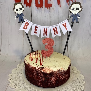 Op de afbeelding: Rode velvet cake met witte frosting en rode sprinkles. De cake is versierd met een rood glitter cijfer 3, een banner met de tekst "BENNY" en twee cartoon figuren van een man met een mes.