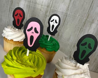 May include: Cupcakes with colorful frosting and Scream mask toppers. The cupcakes have white, green, and yellow frosting. The Scream mask toppers are red, pink, white, and green with black outlines.