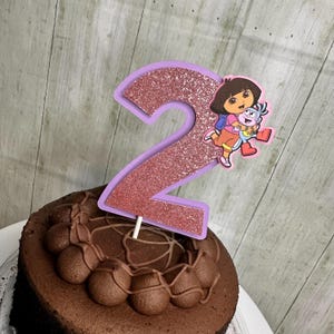 May include: A cake topper featuring a large number two with a rose gold glitter finish and a light purple border. Attached to the side of the number is a colourful illustration of the cartoon character Dora the Explorer hugging her friend Boots.