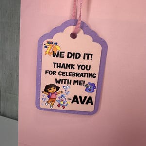 Pode incluir: Uma etiqueta de presente lavanda com uma borda ondulada e uma fita rosa. A etiqueta apresenta o texto "WE DID IT! THANK YOU FOR CELEBRATING WITH ME! -AVA" junto com ilustrações de personagens de desenhos animados.