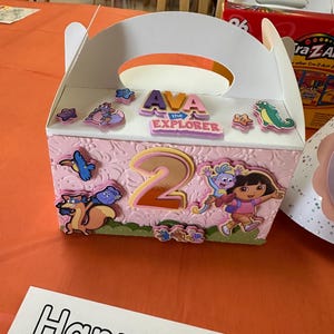 Könnte beinhalten: Eine rosa und weiße Papp-Geschenkbox mit Griff. Die Box ist mit einem Dora the Explorer-Charakter, einer Nummer 2 und den Worten "AVA the EXPLORER" verziert.