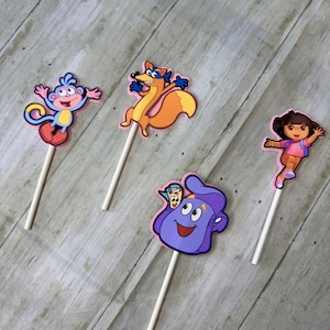Puede incluir: Cuatro adornos coloridos para pasteles con personajes de dibujos animados. Los adornos incluyen un mono, un zorro, una niña y una mochila morada, cada uno unido a un palito blanco. Los personajes están delineados en rosa.