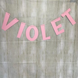 Könnte beinhalten: Ein rosa Banner mit dem Namen "VIOLET" hängt vor einem hellen Hintergrund in Holzoptik. Die Buchstaben sind aus rosa Papier ausgeschnitten und an einer dünnen, silbernen Schnur befestigt. Das Banner ist ein dekoratives Element.