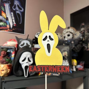 Può includere: Un topper per torta a forma di coniglio giallo con una maschera di fantasma bianca e il testo "EASTERWEEN" in glitter rosso. Il topper è davanti a uno scaffale con vari personaggi e oggetti di scena di film horror.