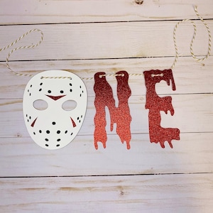 May include: A white mask with black dots and red accents hangs next to the word "ONE" in red glitter letters. The banner is strung on a thin, off-white string, perfect for a first birthday party.