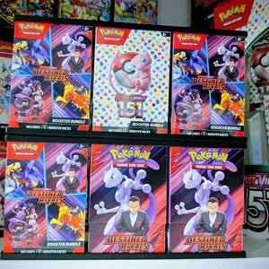 May include: A display of Pokémon trading card game booster bundles and packs. The boxes feature various Pokémon characters and the text "Destined Rivals" and "Scarlet & Violet 151". The color scheme includes red, purple, and white.