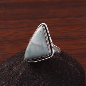 May include: A silver ring featuring a triangular, pale blue gemstone. The gemstone is set in a silver bezel, and the ring is displayed on a dark, textured surface. The ring's design is simple yet elegant, suitable for everyday wear.