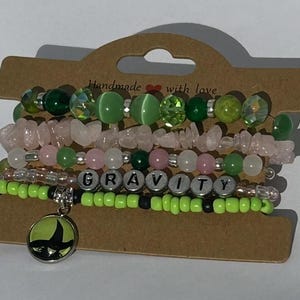 May include: A collection of beaded bracelets in shades of green, pink, and clear. One bracelet spells out "GRAVITY" with letter beads. A charm with a green and black design hangs from one bracelet. The bracelets are displayed on a brown card.