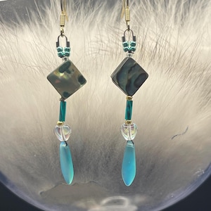 Può includere: Un paio di orecchini pendenti con ganci color oro. Ogni orecchino presenta una perla quadrata iridescente, perline turchesi, perline trasparenti a forma di cuore e una perla turchese a forma di goccia. Gli orecchini sono esposti in un bicchiere trasparente.