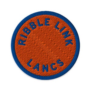 May include: A circular embroidered patch featuring a vibrant orange center with a textured stitch pattern. The text RIBBLE LINK LANCS is stitched in bold blue thread along the perimeter, framed by a thick blue border around the circular edge.