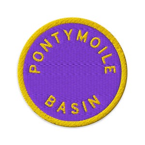 Puede incluir: Un parche bordado circular con un centro de color morado vibrante y un borde grueso de color amarillo. El texto PONTYMOILE BASIN está bordado en letras mayúsculas amarillas, siguiendo la curva circular del borde sobre el fondo morado.