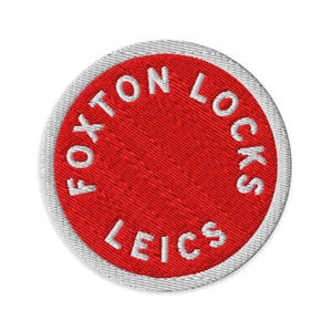 Pode incluir: Um emblema bordado circular com um centro vermelho vibrante e uma borda branca. O texto FOXTON LOCKS LEICS está bordado em letras maiúsculas brancas ao longo da borda interna do círculo vermelho, seguindo a forma circular do design.
