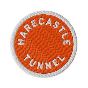Pode incluir: Um emblema bordado circular com um fundo laranja vibrante e uma borda branca. O texto "HARECASTLE TUNNEL" está bordado em letras maiúsculas brancas, curvando-se ao longo da borda interna do emblema em uma disposição circular.