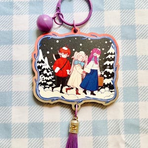 May include: A purple star-shaped keyring featuring a rectangular charm with an illustration of three figures walking through a snowy forest. The charm includes a purple metal ring, a small purple bell, and a long purple tassel hanging from the bottom.