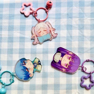May include: Three acrylic keychains featuring anime-style character heads. One has pink hair and a blue bow, one has blue hair, and one has purple hair. Each keychain includes a matching coloured metal ring, a star-shaped charm, and a small round bell.