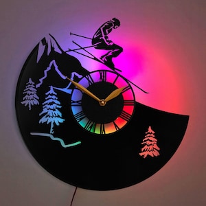 May include: A black vinyl record clock featuring a skier silhouette and a rainbow clock face. The clock is illuminated with multi-coloured LED lights.