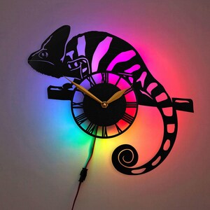 May include: Black chameleon-shaped wall clock with a circular clock face featuring Roman numerals and gold-colored hands. The clock is illuminated with a vibrant array of colors, including pink, purple, blue, green, and red.