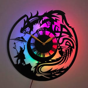 May include: A black and white silhouette of a dragon and a wizard holding a staff, with a clock face in the center. The clock face has a rainbow-colored background and gold hands. The clock is a wall clock with a light-up feature.