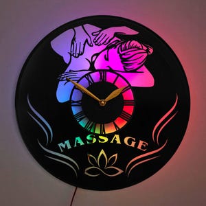May include: A black vinyl record clock with a colorful LED light display. The clock features a silhouette of a person receiving a massage with the word "MASSAGE" printed below. A lotus flower design is also visible.