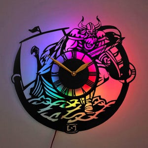 May include: A black and white wall clock with a Viking warrior design. The clock has a rainbow LED light that illuminates the design. The clock is made of wood and has a black background.