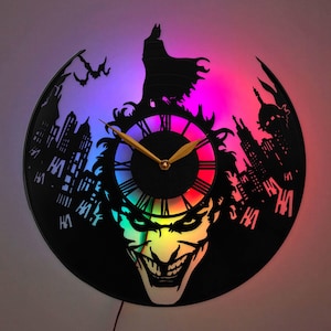 May include: A circular wall clock crafted from a vinyl record featuring a cutout of a caped figure standing above a grinning face. The design includes city silhouettes, bats, and the text HA. It is illuminated by a vibrant blue and pink LED light.