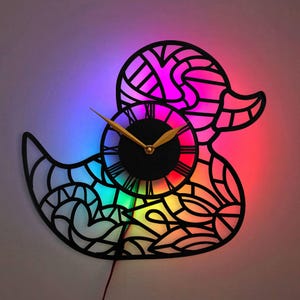 May include: A black, laser-cut, duck-shaped wall clock with a rainbow-coloured LED light behind it. The clock has Roman numerals and gold-coloured hands.