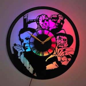 May include: A black and white clock with a rainbow-coloured clock face. The clock features silhouettes of three horror movie characters: Michael Myers, Jason Voorhees, and Freddy Krueger.