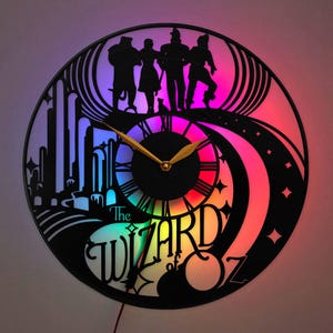 May include: A circular wall clock crafted from a black vinyl record featuring a silhouette cutout of characters from The Wizard of Oz. The design includes the Emerald City, a winding road, and the text The Wizard of Oz, illuminated by colorful LED lights.