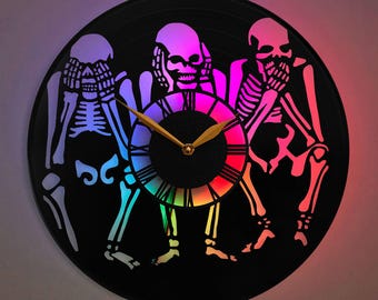 May include: A black vinyl record clock featuring three skeletons in a circle, each covering their eyes, ears, and mouth. The clock face is rainbow coloured and has gold hands.