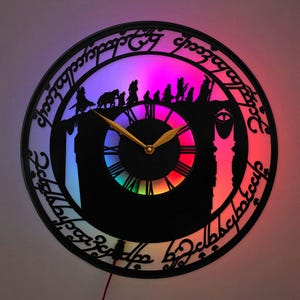 May include: A circular wall clock featuring a black silhouette cutout of a group of figures walking along a ridge, with the Eye of Sauron on the right. The outer ring displays Elvish script. The clock face is illuminated by a vibrant, multi-colored LED light.