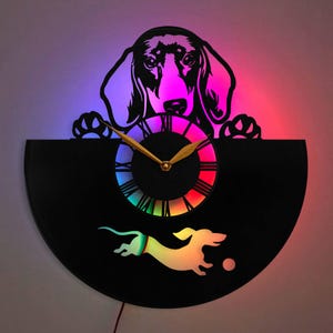 May include: A black vinyl record clock with a dachshund silhouette peeking over the top. The clock face is a rainbow gradient with gold hands. The dachshund is also depicted running with a ball in its mouth.