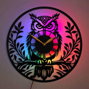 May include: A black owl clock with a rainbow-coloured LED light behind it. The owl is perched on a branch with leaves. The clock face is black with gold hands and Roman numerals.
