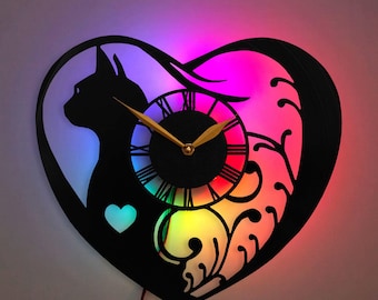 May include: A black silhouette of a cat with a heart on its chest is on the left side of a heart-shaped clock. The clock face is black with gold hands and Roman numerals. The background is a rainbow gradient with a floral design.