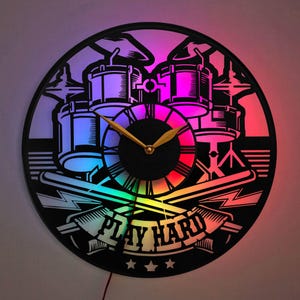 Op de afbeelding: Een zwart-witte drumset met een klok en de woorden "Play Hard" in een banner. De klok heeft gouden wijzers en een regenboogkleurig licht erachter.