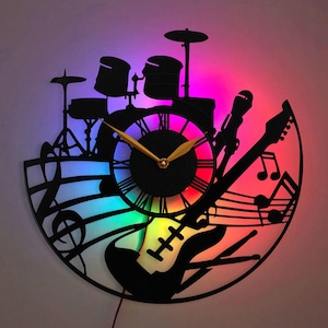 Op de afbeelding: Een zwarte metalen wandklok met een gitaar, drums en muzieknoten. De wijzerplaat van de klok heeft Romeinse cijfers en een gouden uur- en minutenwijzer. De klok heeft een regenboogkleurig LED-licht erachter.