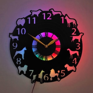 Könnte beinhalten: Eine schwarze Schallplattenuhr mit einem Regenbogen-LED-Lichtdisplay. Die Uhr zeigt für jede Stundenmarkierung eine Silhouette einer Hunderasse. Die Zeiger der Uhr sind golden.