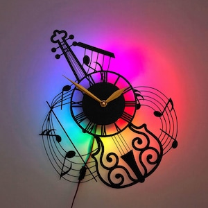Op de afbeelding: Een zwarte metalen wandklok in de vorm van een viool met muzieknoten en een regenboogkleurig LED-licht. De wijzerplaat van de klok heeft Romeinse cijfers en goudkleurige wijzers.
