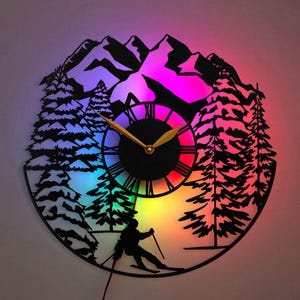 May include: A black and white silhouette of a mountain scene with a skier, trees, and a clock face with gold hands. The clock face is illuminated with a rainbow of colors.