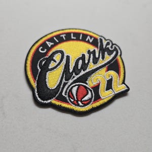 May include: A black and yellow embroidered patch with the name "Caitlin Clark" in a white cursive font and the number "22" in red. A basketball is in the center of the patch.