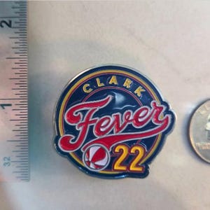 Puede incluir: Un pin circular azul y amarillo con el texto "Clark Fever 22" en letras rojas y blancas. Un gr&aacute;fico de baloncesto est&aacute; en el centro del pin.