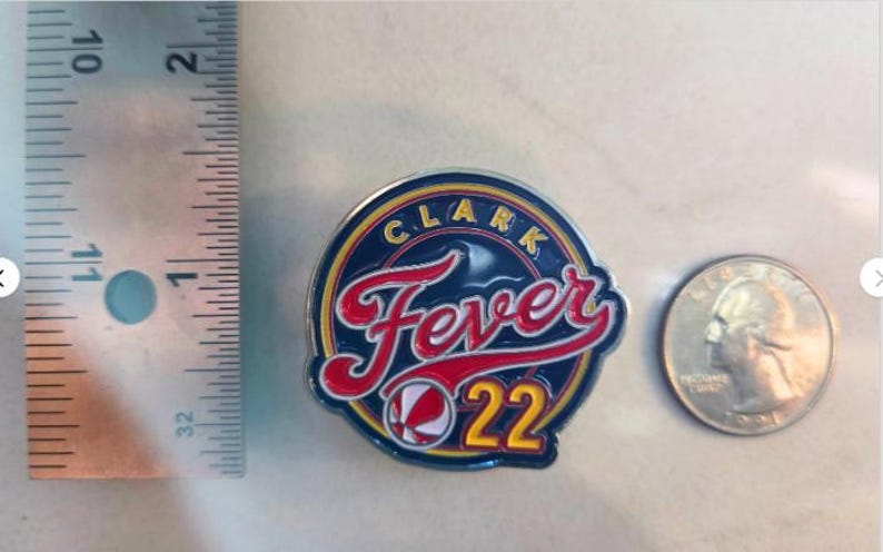 Puede incluir: Un pin circular azul y amarillo con el texto "Clark Fever 22" en letras rojas y blancas. Un gr&aacute;fico de baloncesto est&aacute; en el centro del pin.