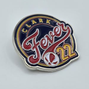 Puede incluir: Un pin de metal plateado con un dise&ntilde;o azul, rojo y amarillo. El pin presenta el texto "Fever 21" en una fuente estilizada y un gr&aacute;fico de baloncesto. El anillo exterior del pin tiene el texto "Clark" en amarillo.