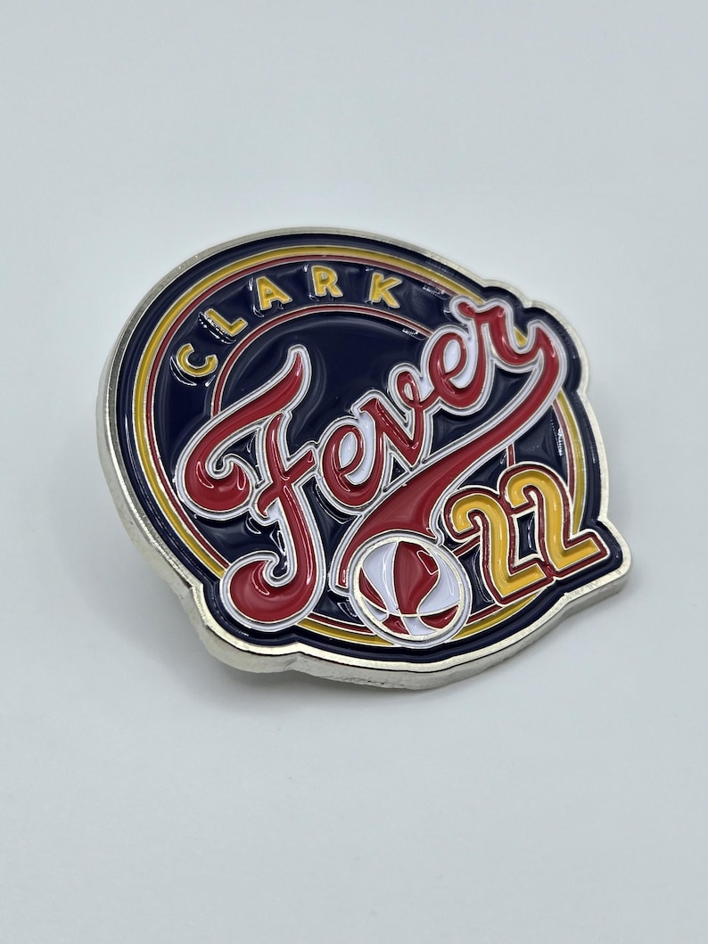 Puede incluir: Un pin de metal plateado con un dise&ntilde;o azul, rojo y amarillo. El pin presenta el texto "Fever 21" en una fuente estilizada y un gr&aacute;fico de baloncesto. El anillo exterior del pin tiene el texto "Clark" en amarillo.