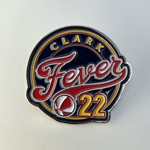 Puede incluir: Un pin circular azul y dorado con el texto "Clark Fever 22" en una fuente de letra blanca y roja. Un gr&aacute;fico de baloncesto est&aacute; en el centro del pin.